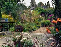 An autumn gravel garden with large informal borders of perennials and grasses with yew and box structure. In the foreground orange cactus Dahlia, pink Salvia 'Bethelii', Pennisetum Rubrum and containers of scented pelargonium.