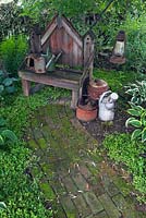 Brick path covered with green Bryophyta - Moss and old wooden sitting bench decorated with antique watering cans in backyard rustic garden in summer, Quebec, Canada