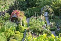 View from above of rural garden with arbour covered with rambling rose 'Excelsa', box balls, vegetable patch, gravel paths and a clematis arch over a wooden garden gate.