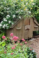 Wooden chicken house covered with a climbing white Rose, Centranthus ruber in the foreground