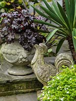 Weathered stone cockerel sits amidst pots of bacopa, heuchera and cordyline.