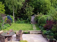 A family garden with stone patio with seating and dining area. Two steps lead up to the lawn. Seen from upstairs window. Wicker furniture 