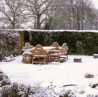 Lutyens style bench and table with dusting of snow on stone terrace sheltered by wall and hedge.
