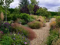A gravel path lined by double mixed borders. Planting includes Leucanthemum, Achillea, Sedum, Eupatorium, Echinacea, Monarda, Guara and Rudbeckia with Stipa and Calamagrostis