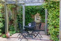 A stone and brick patio seating area covered by a wooden pergola. On the back wall a ceramic water feature from Lucy Smith Garden Sculptures
