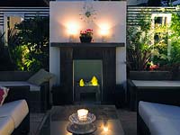 Outdoor room at night with sofas by gas effect fire and fireplace. Backlit raised beds to each side, behind built-in seating.