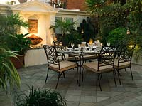 Walled outdoor room at night with illuminated wall fountain mask trickling water into raised pool flanked by pots of palms and arum lilies. Table laid for dinner.
