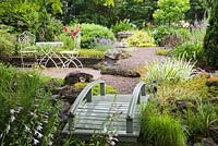 Grey painted wooden footbridge bordered by Acorus gramineus 'Variegatus - Ornamental Grass plants leading to a white cast-iron metal table and chairs in backyard country garden in summer, Quebec, Canada
