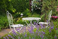 Lavandula angustifolia 'True Blue' - Lavender, red Lilium Asiatic 'America' - Liliy flowers  and white cast-iron metal table and chairs,  white Clematis 'Huldine' growing on brown wooden pergola in the background in backyard country garden in summer, Quebec, Canada