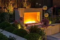 Town garden designed by Kate Gould, lit at night. Flames from an open gas fireplace illuminate the sunken terrace which is edged in beds of box balls interspersed with purple and white allium. Back boundary is planted with tall golden bamboo.