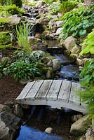 Creek with cascading waterfalls and grey wooden footbridge bordered by Hosta plants in backyard garden in summer, Quebec, Canada
