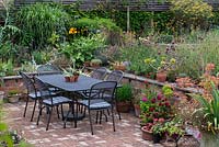 An outdoor dining table and chairs on the brick terrace, with retaining wall, mixed perennials and container planting.
 