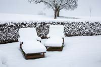 Snow on wooden deck chairs, a hedge and the fruit trees in the landscape