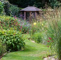 A grass path leads to a summerhouse  between herbaceous borders planted with Monarda, Persicaria, Dahlia, Verbena bonariensis with Stipa and Calamagrostis grasses.