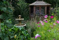 A stone water feature and wooden summerhouse amongst a border planted with Cosmos, Verbena bonariensis and Salvia.