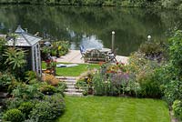 Bird's eye view of summerhouse by bed of catmint, coneflower, annual poppies and oriental lilies. Lawn leads to riverside deck edged in pots and beds of oriental lilies, daylilies, Verbena bonariensis, annual poppies, agapanthus and kniphofia.