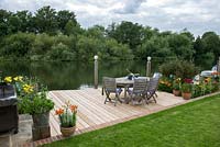 Riverside deck with barbecue, dining table and chairs, is edged in pots and beds of oriental lilies, daylilies, Verbena bonariensis, annual poppies, agapanthus, dogwood and kniphofia.