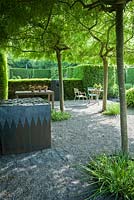 Graveled courtyard area beside the studio with four weeping ash, Fraxinus excelsior 'Pendula' forming a shady canopy, steel and lead container, filled with an arrangement of stone setts and a seating area surrounded by yew hedging. 