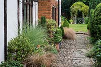 A stone path with terracotta containers planted with ornamental grasses and annuals. In the distance, a clematis covered gazebo with seating area. Southend Farm.