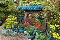 Oriental raku-tiled mirrored wall by artist owner, edged in bamboo, Nandina domestica and ivy. Surrounded by pots of tulips, self-seeded forget-me-not and euphorbia.