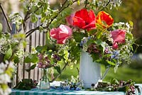 Display of spring flowers in enamel jug that includes tulips, Prunus padus - bird cherry, Myosotis arvensis, Allaria petriolata - garlic mustard, Lamium orvala. Flowering pear tree behind - Pyrus 'Williams'.