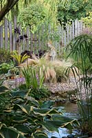 A gravel garden with Stipa grasses surrounding a stone statue behind a shady pond.
