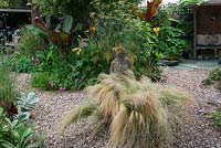 A gravel garden with Stipa grasses surrounding a stone statue in front a border with tropical foliage plants.