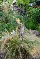 A gravel garden with Stipa grasses surrounding a stone statue in front a border with tropical foliage plants and perennials.