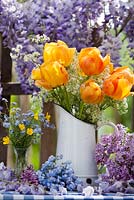 Outdoor spring display - tulips and cow parsley in jug, forget me nots and buttercups. Wisteria climbing against wooden fence.