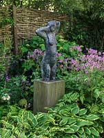 Sculpture of woman on high wooden plinth rises above hosta, rose, hydrangea and Geranium maderense.