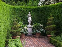 Classical statue on plinth in formal brick courtyard enclosed in curving yew hedge. Pots of box spirals and Datura.