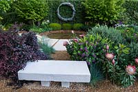 The Time In Between by Husqvarna and Gardena. View of  concrete bench, stone water feature and circular copper sculpture surrounded by Protea cynaroides 'Little Prince', Alternanthera dentata, Polygala myrtifolia, Festuca Glauca, Westringia fruticosa 