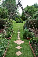 Rustic hazel arch frames garden. Path through wooden edged vegetable and herb beds with diamond shaped stepping stones 