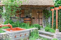 View of whole garden with thatched stone shed. Brewing barrel, water feature featuring beer taps. Clipped balls of Buxus. Brewers Yard by Welcome to Yorkshire, RHS Chelsea Flower Show 2015 