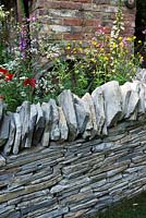 The Old Forge Garden for Motor Neurone Disease Association. RHS Chelsea Flower Show 2015. Natural slate border wall surrounded by wild flowers Digitalis purpurea, Papaver rhoeas, Anthriscus sylvestris, Ranunculus acris, against brick wall. 