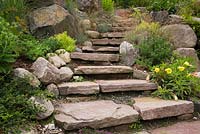 Natural stone steps borderd by perennial plants and shrubs, yellow Lilium - Lily flowers in backyard garden bordered by perennial shrubs and flowers in summer