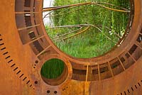 A rusted corten steel oculus and warped steel rods depicting the trajectory of light flow through the garden planted with foliage plants including Phyllostachys and grasses. Dark Matter Garden for the National Schools' Observatory. RHS Chelsea Flower Show 2015. 