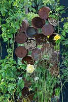 The  Great Chelsea Garden Challenge. Rusted cans make decorative divider, with sawn-off canes for insect habitats. Rosa 'Golden Gate' and grasses in foreground.