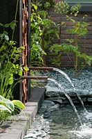 Ebb and Flow of water through copper piping with Angelica sylvestris 'Ebony' growing in the gravel and Hosta sieboldiana 'Frances Williams' by the water.  RHS Chelsea Flower Show, 2015