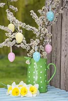Decorative eggs hanging from blossoming spring foliage in a green polkadot jug, accompanied with Daffodils