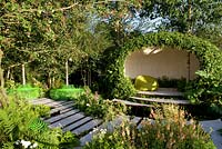 White stone paving through shady woodland-edge garden with pod structure seating area. Green-painted seats around stem of birch trees.  The Macmillan Legacy Garden RHS Hampton Court Palace Flower Show 2015. Sponsor Macmillan Cancer Support 