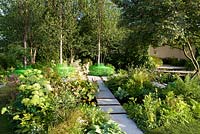  White stone paving through shady woodland-edge garden with pod structure seating area. Three birch trees with green-painted seats. The Macmillan Legacy Garden Sponsor Macmillan Cancer Support. RHS Hampton Court Palace Flower Show 2015
