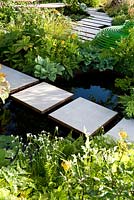 White slab stepping stones over black-water pool with naturalistic green, shade tolerant planting. The Macmillan Legacy Garden Sponsor Macmillan Cancer Support. RHS Hampton Court Palace Flower Show 2015