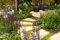 Curving path of stone slabs leading to circular seating area and raised water feature surrounded by flower beds planted with purple blue and white perennials and grasses. Agapanthus africanus Lupinus Salvia nemorosa Cardonna Chamaemelum nobile Pennisetum orientalis Digitalis Camelot White Salvia officinalis Purpurascens Thymus serphyllum Geranium Rozanne and Verbena rigida  in The Wellbeing of Women Garden at RHS Hampton Court Palace Flower Show 2015 