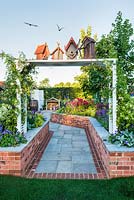 Just Retirement: A Garden For Every Retiree, view of white pergola with birdhouses and birds at the entrance to rural garden. Designer: Tracy Foster Sponsor: Just Retirement Ltd