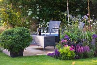 A garden by association, view of seating area with synthetic wicker armchairs and a table surrounded by Betula Jacquemontii, Anthriscus sylvestris, flowerbed with Cosmos, Veronica longifolia 'Charlotte', Nepeta x faassenii 'Blue Wonder', Sambucus nigra porphyrophylla 'Eva' and pot with Pittosporum Tobira - Designer: Tina Vallis, MSGD

