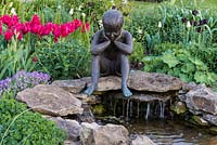 Figurine of a contemplative child, sculpted by Jenny Wynn Jones, overlooks a stone pool. Behind, red Tulipa 'Pimpernel'.