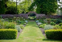 View through walled garden to the Spider Gate, with woodland beyond. Box - Buxus sempervirens edging and growing herbaceous borders in spring in foreground
