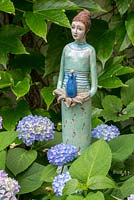 Female ceramic sculpture holding a stylized bell flower next to Hydrangea macrophylla  'Endless Summer'