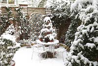 Courtyard with seating area and pots in winter. Picea glauca var. albertiana 'Conica' in pot on table with decorations made of red ribbon. A mirror trompe l'oeil creates a focal point on a wall of a garage - Welsch Garden, Berlin, Germany